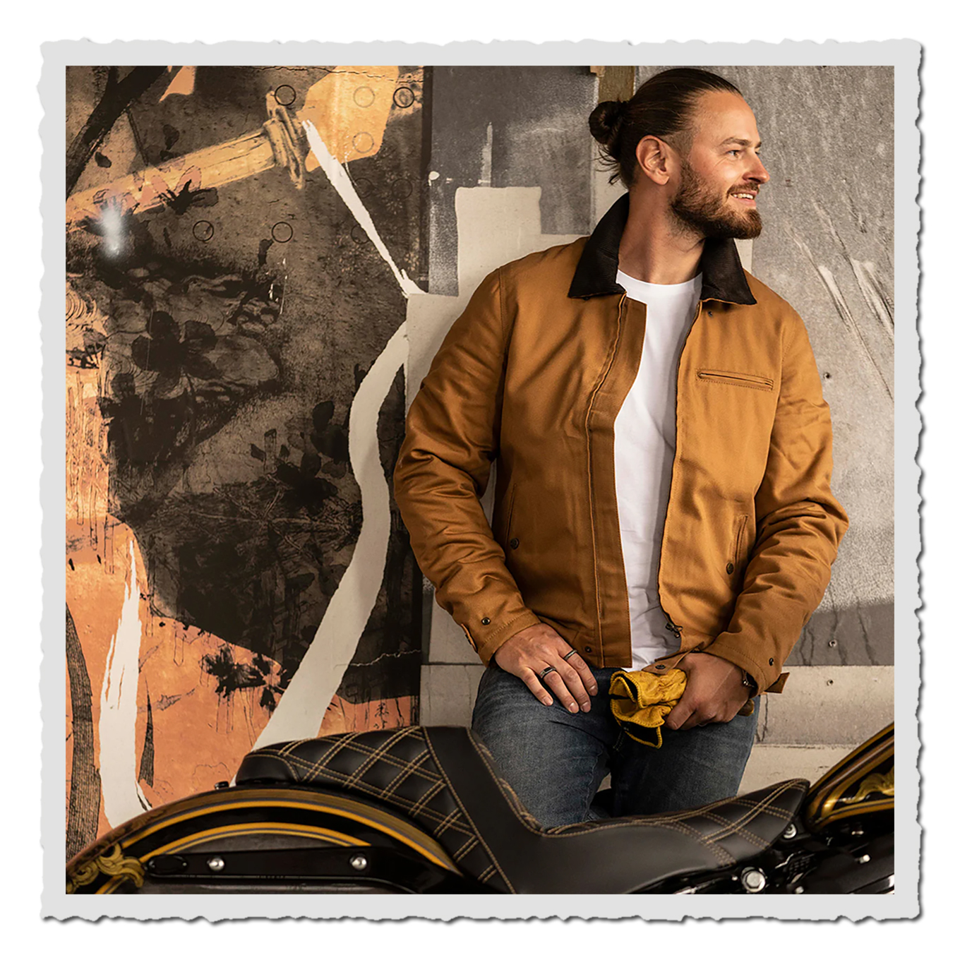 Mann in brauner Jacke und weißem Shirt sitzt neben Motorrad vor gemalter Wand.